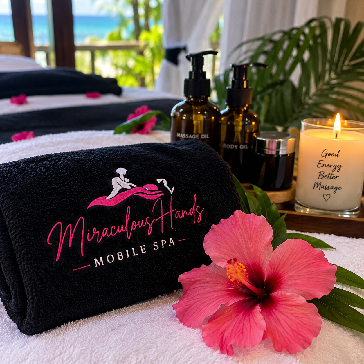 Miraculous Hands Mobile Spa branded black towel with massage oils, a candle and a pink hibiscus by an ocean view